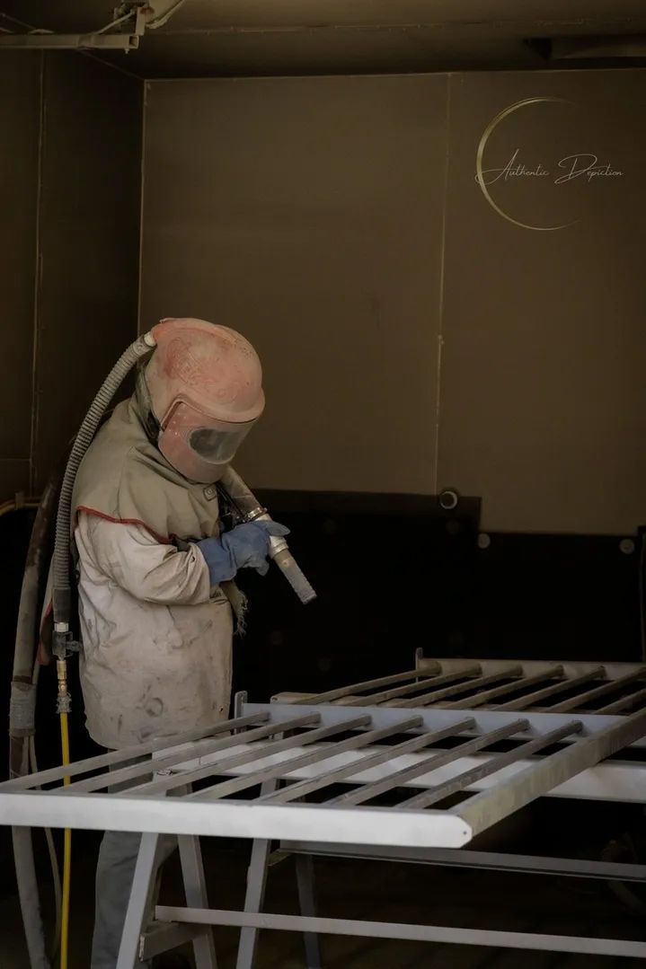 Person sandblasting a metal gate inside a workshop. They wear protective gear and hold a sandblasting gun.