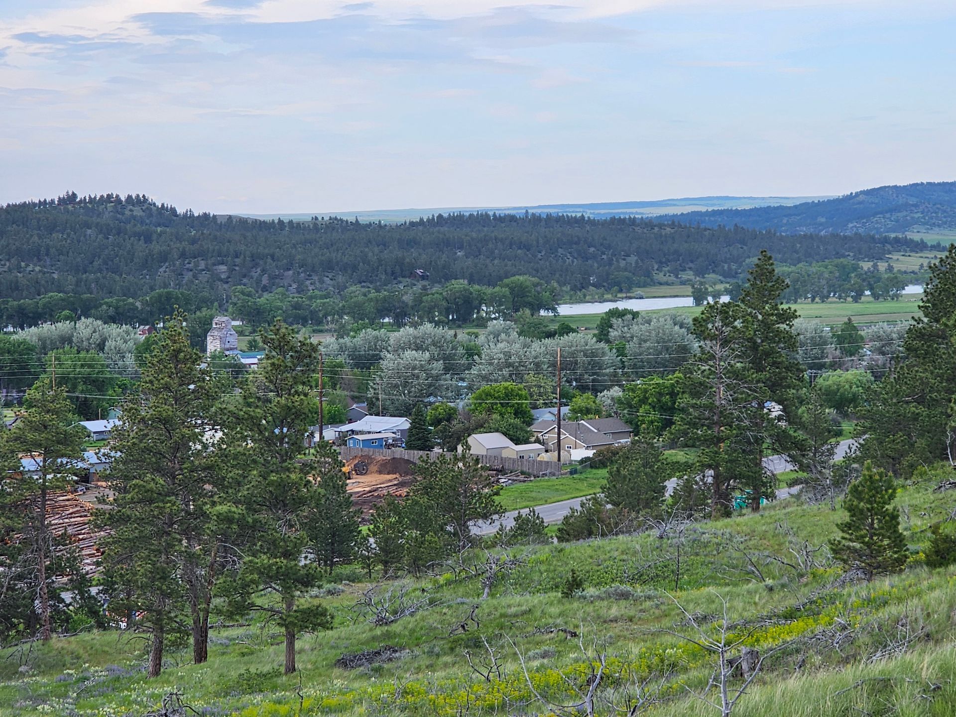 Green hills overlooking a small town with trees, buildings, and a lake under a cloudy sky.