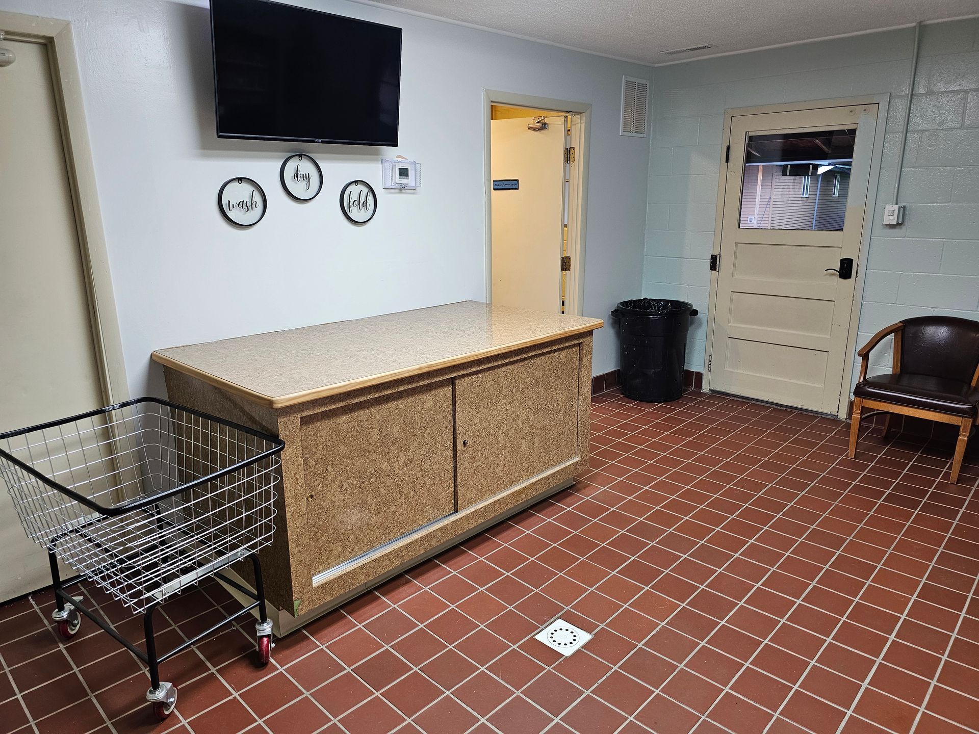 Laundry room with countertop, cart, chair, TV, and trash can. Red tiled floor and tan walls.
