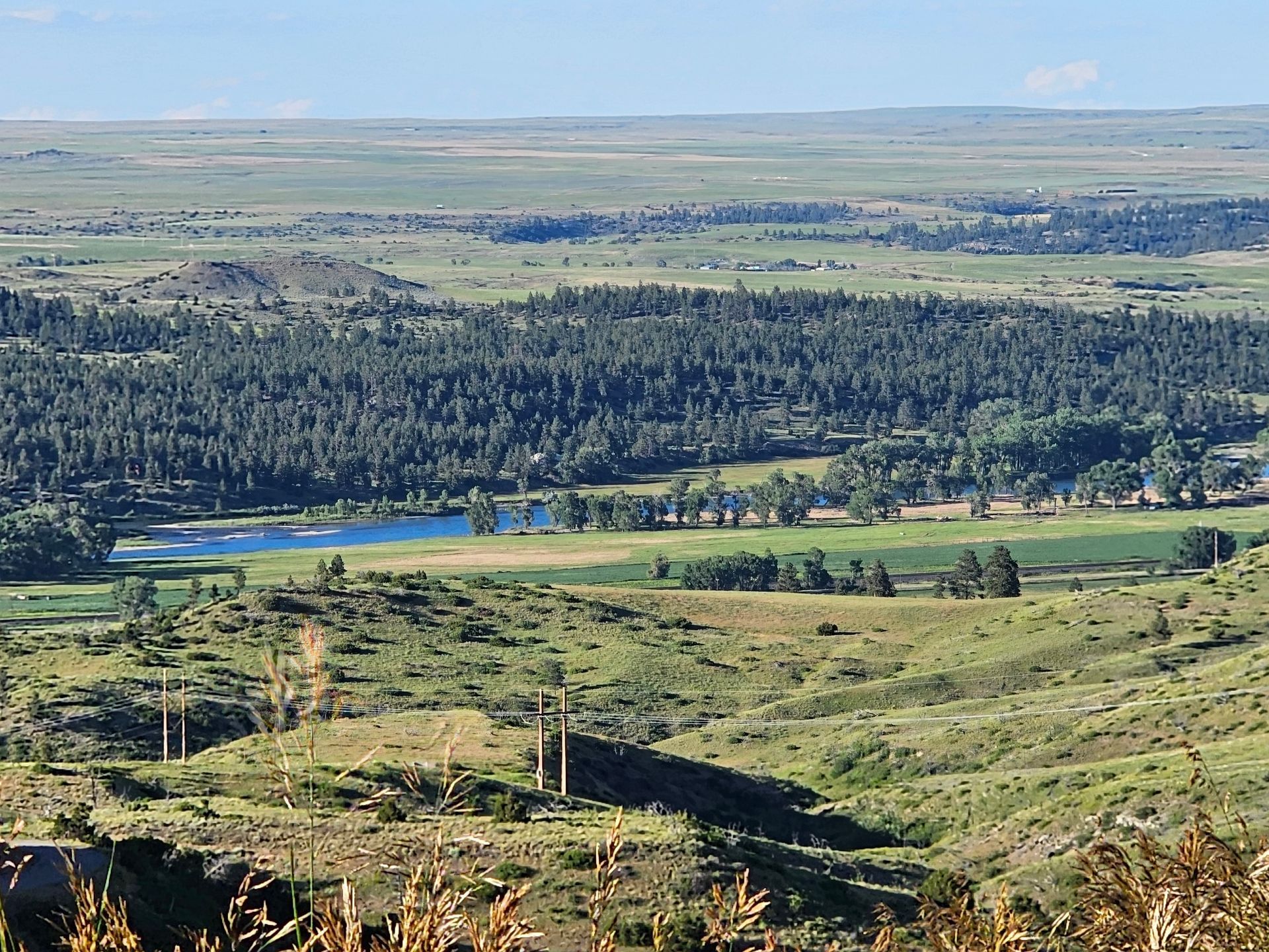Overlook of a valley with trees and a lake, under a vast plain, against a bright, sunny sky.