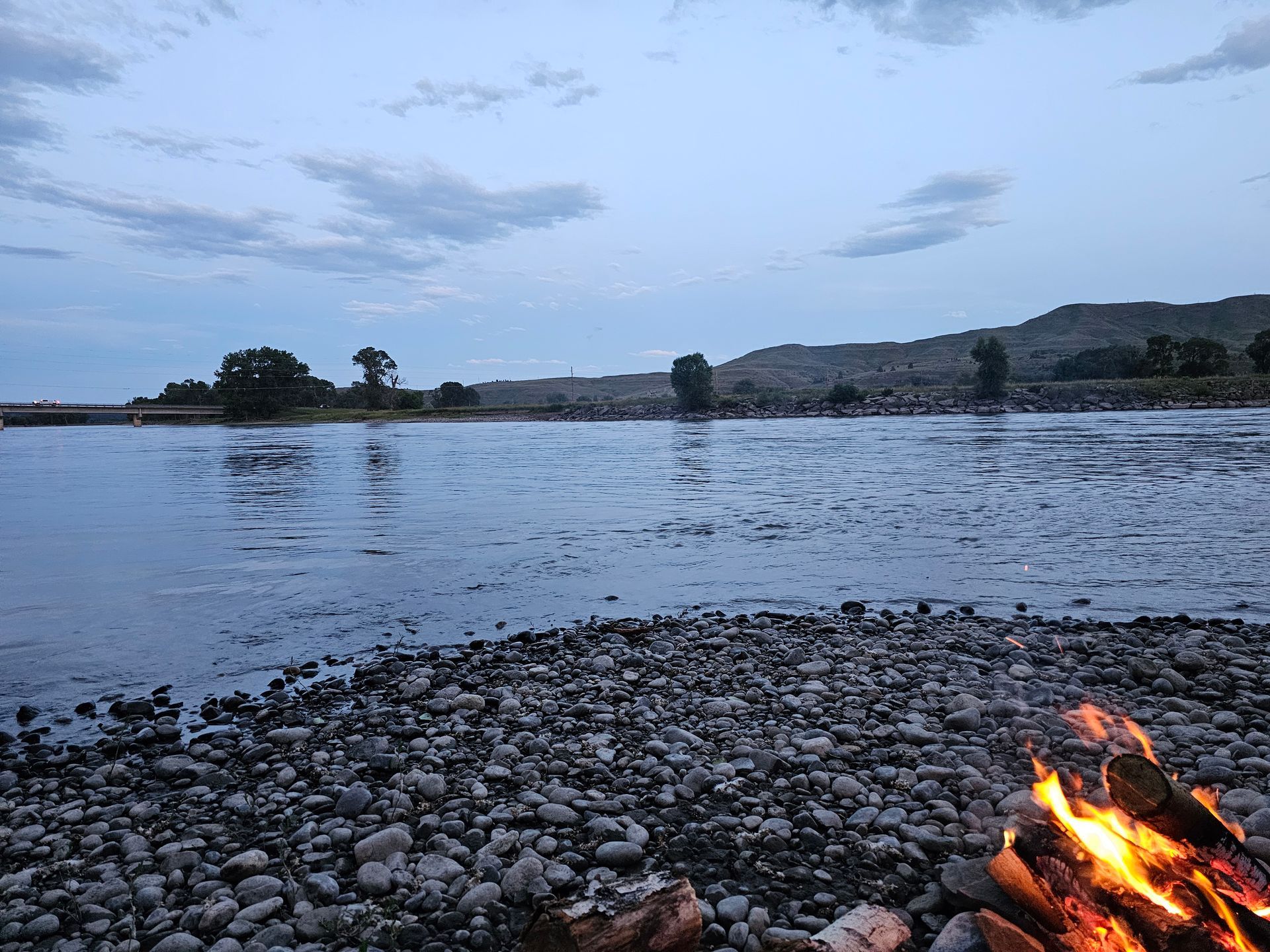 A campfire burns on a rocky shore, overlooking a wide river under a cloudy dusk sky.