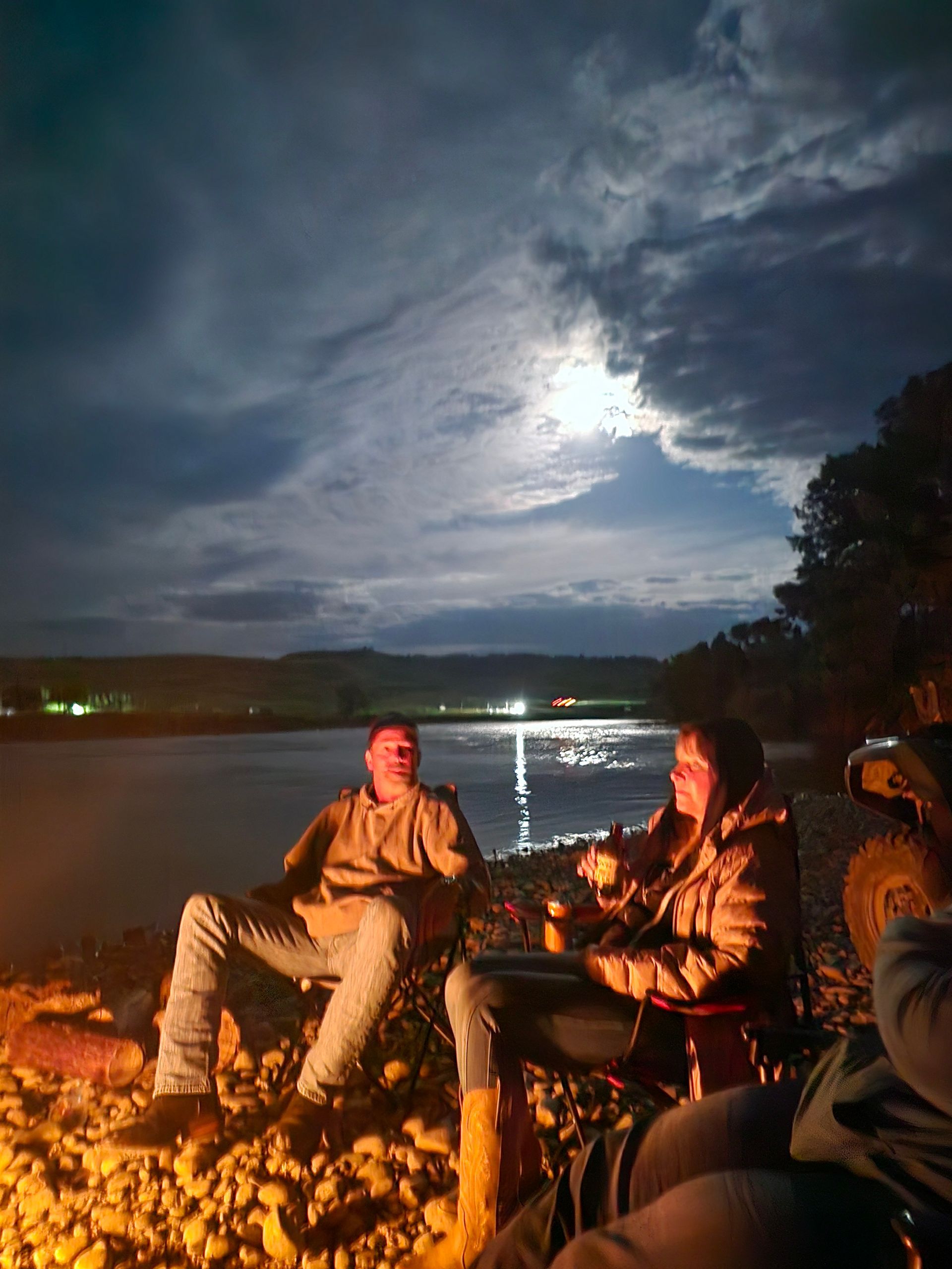 Two people sit by a campfire at night, gazing at the moonlit lake.