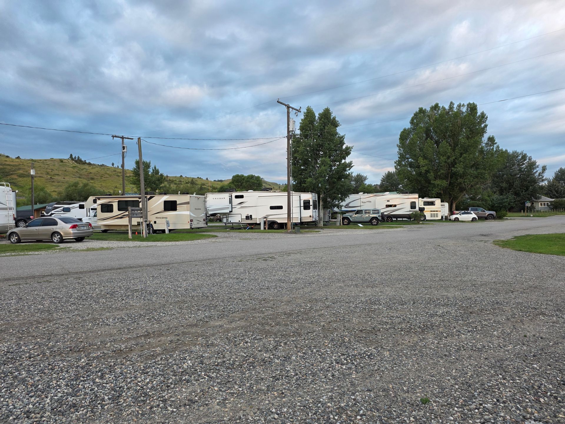 RV park with gravel parking, trailers, trees, and a cloudy sky.
