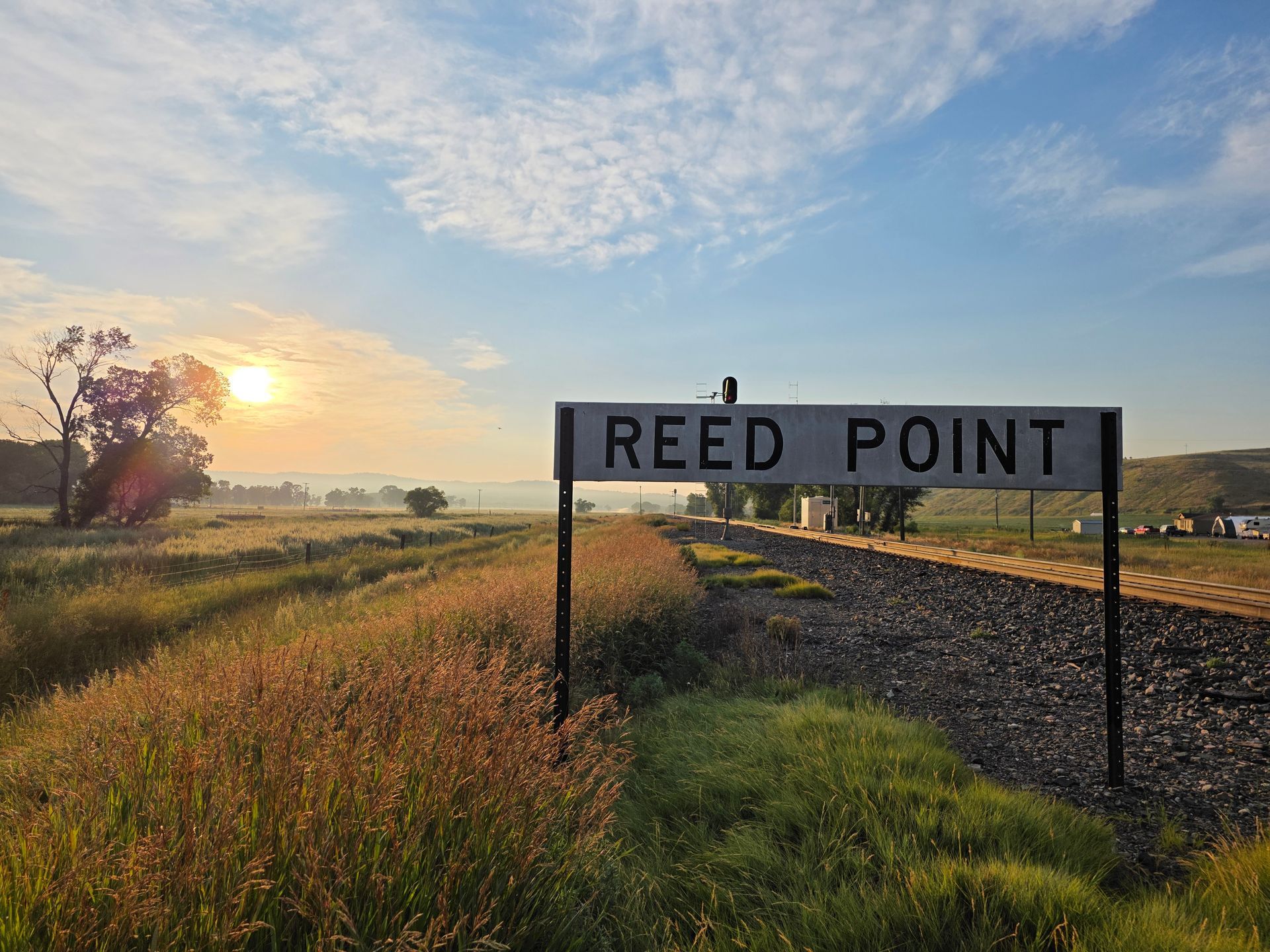 Sign for Reed Point railway station at sunrise, with tracks, grasses, and hazy sky.