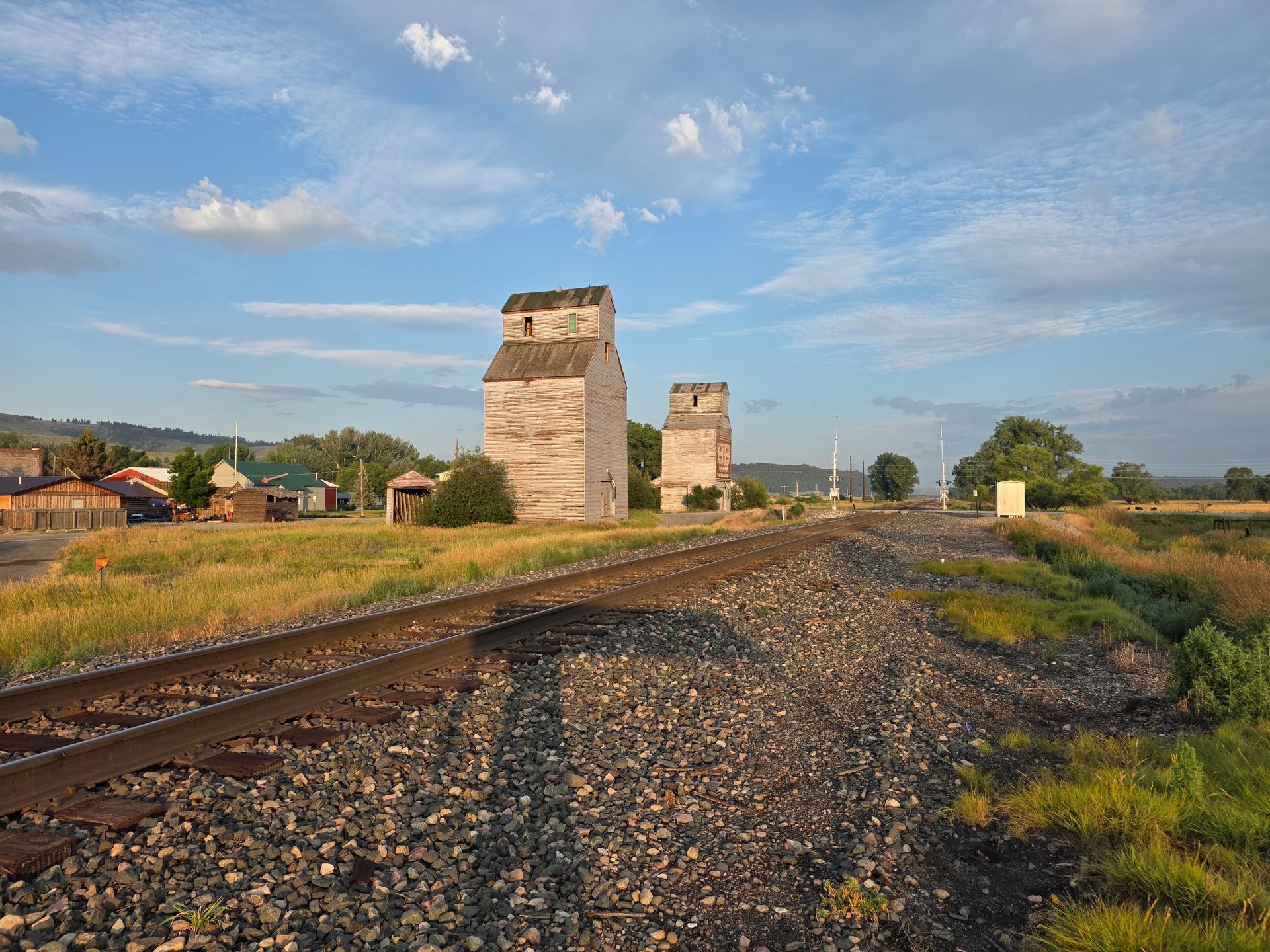 Railroad tracks lead to two weathered grain elevators under a blue sky.