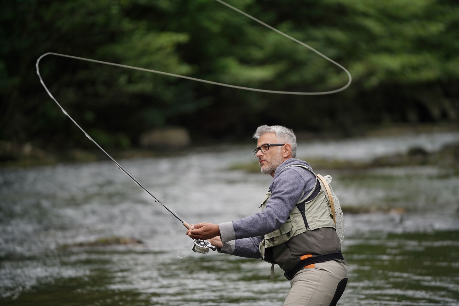 Man fly fishing in a river, casting a line, wearing a vest and glasses.
