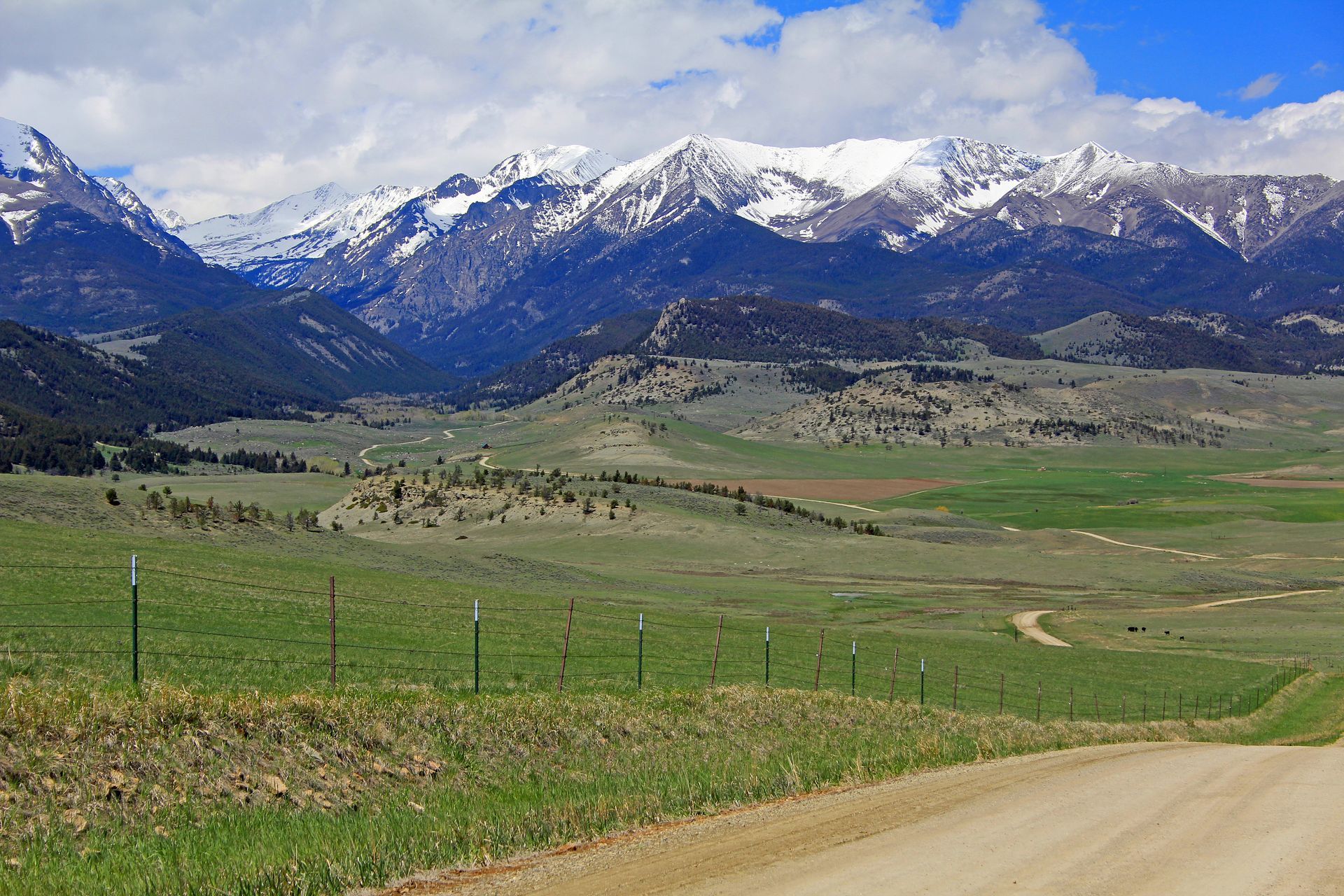 Dirt road leads to a valley with green fields, small hills, and snowy mountain range under a blue sky.