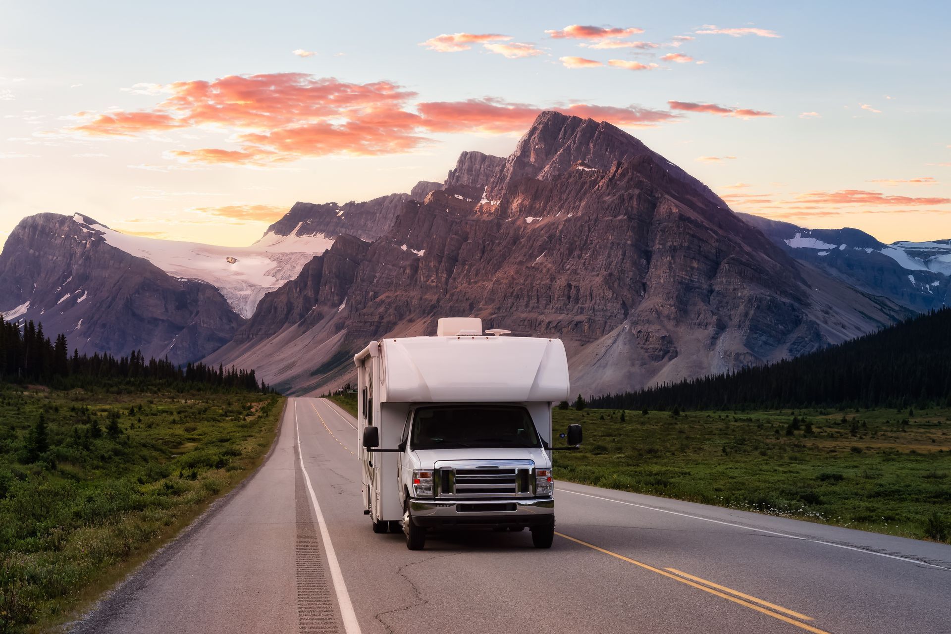 RV driving on a road towards mountains under a sunset sky.
