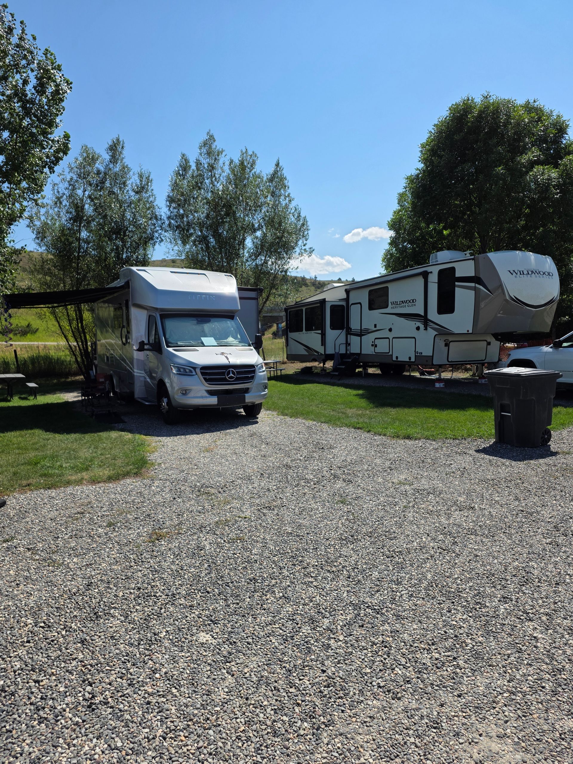 RV camping scene: a white camper van and a larger trailer parked on gravel and grass under a blue sky.