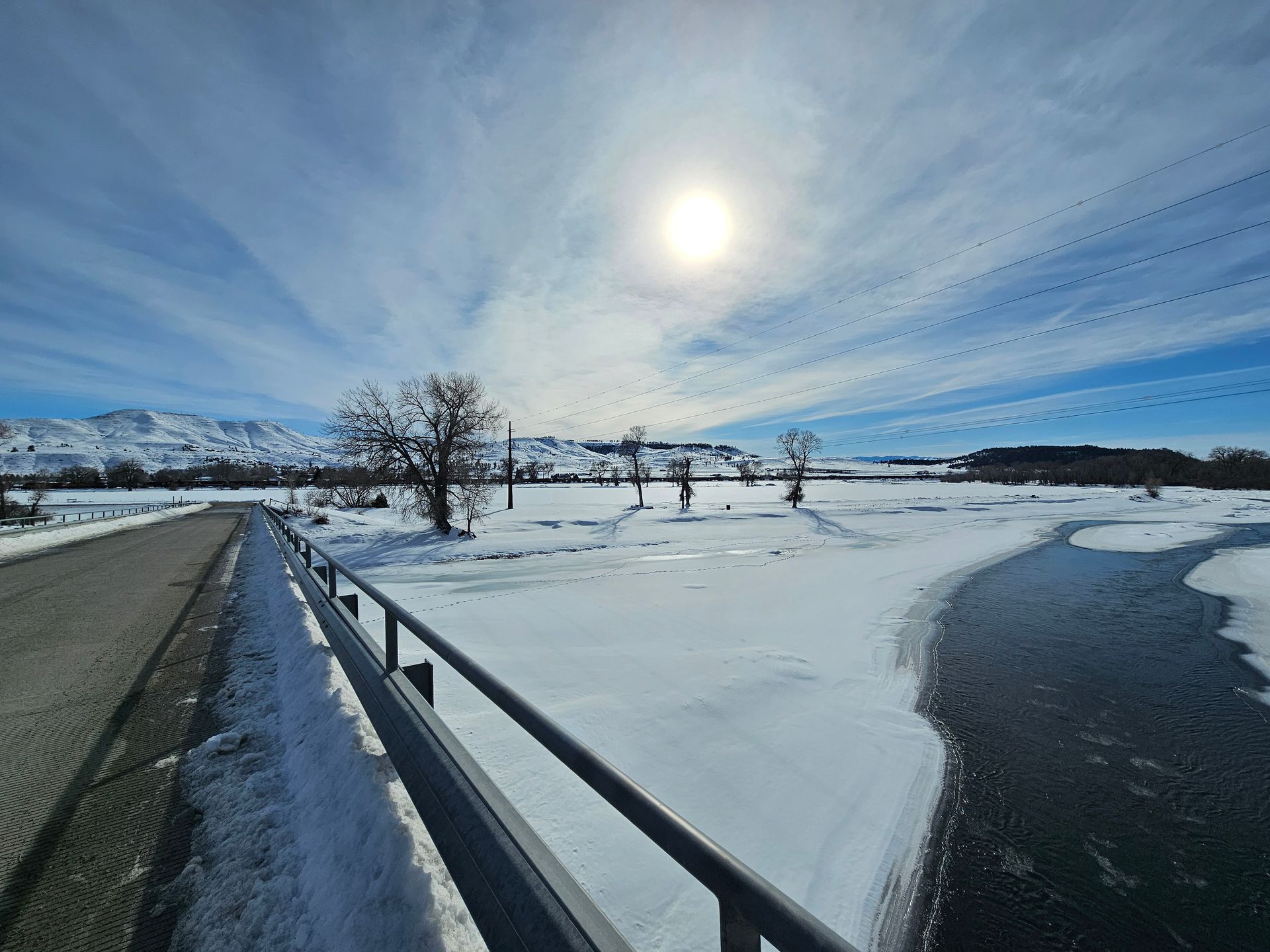 A bridge over a snowy river with the sun shining through the clouds.