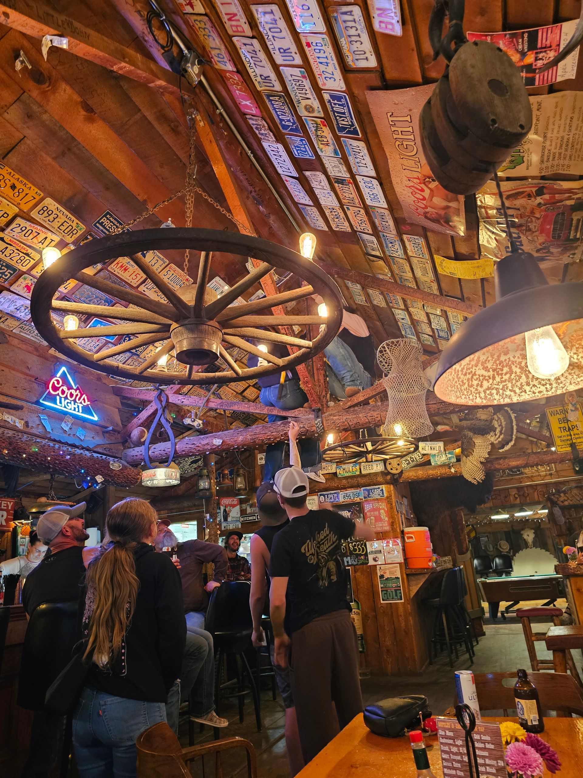 A group of people are sitting at tables in a restaurant with a wagon wheel hanging from the ceiling.