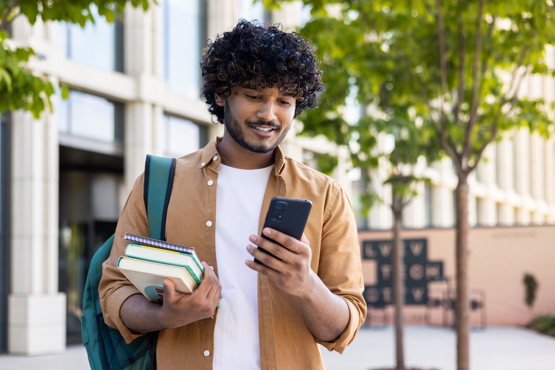 a man holding books and a backpack looks at his phone