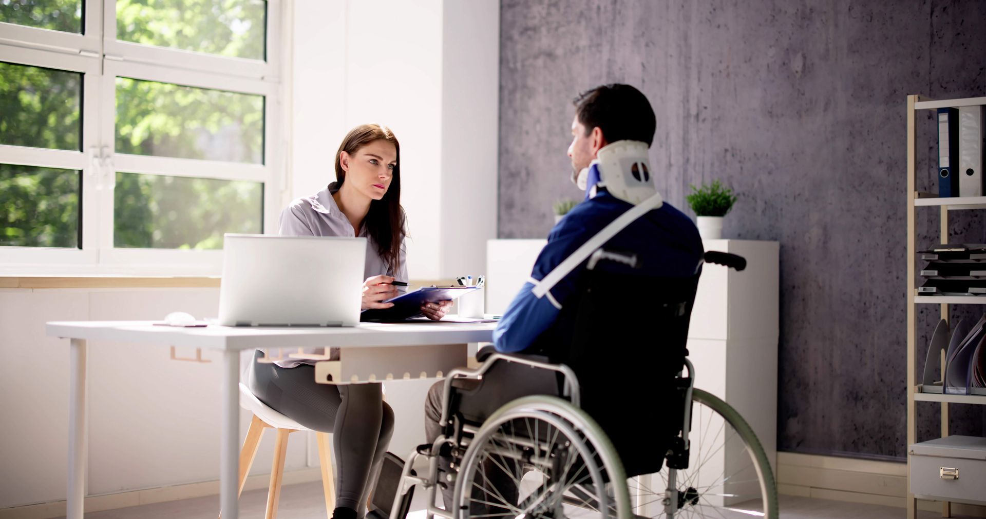 Woman interviewing a man in a wheelchair with a neck brace and arm sling at a desk.