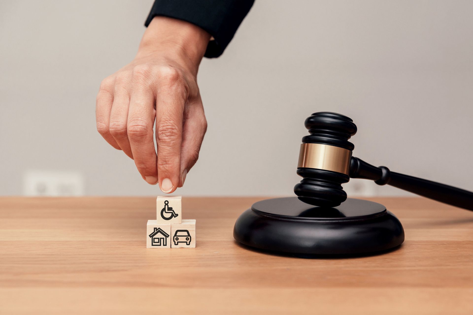 Close-up of a hand stacking blocks with disability, house, and car icons near a judge's gavel. Close-up of a hand stacking blocks with disability, house, and car icons near a judge's gavel.