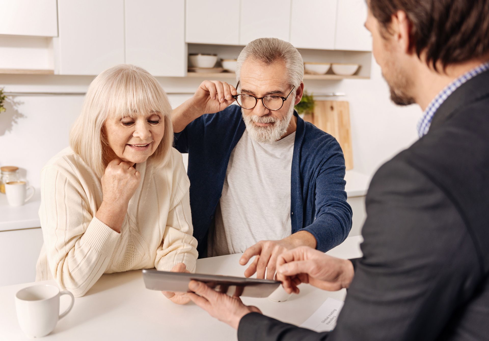 An older couple talks to a lawyer as he shows them something on a tablet, sitting on a living room.