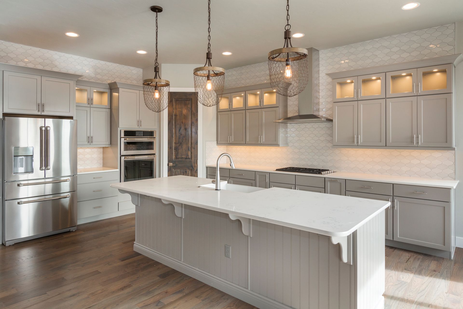 Modern kitchen with gray cabinets, white countertops, and wooden floors. Three pendant lights hang over a large island.