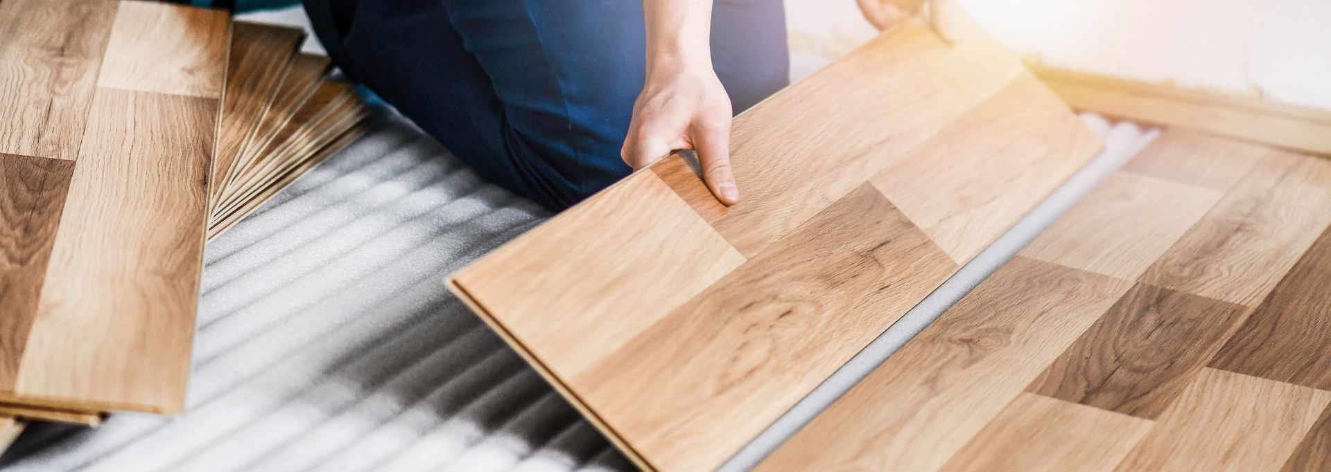 Person installing wood flooring, with hands holding a plank above the floor. Light brown wooden planks and floor.