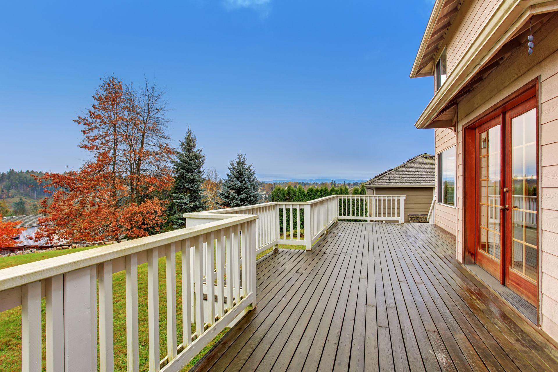 Wooden deck with white railing, overlooking a green lawn and autumn trees under a blue sky.