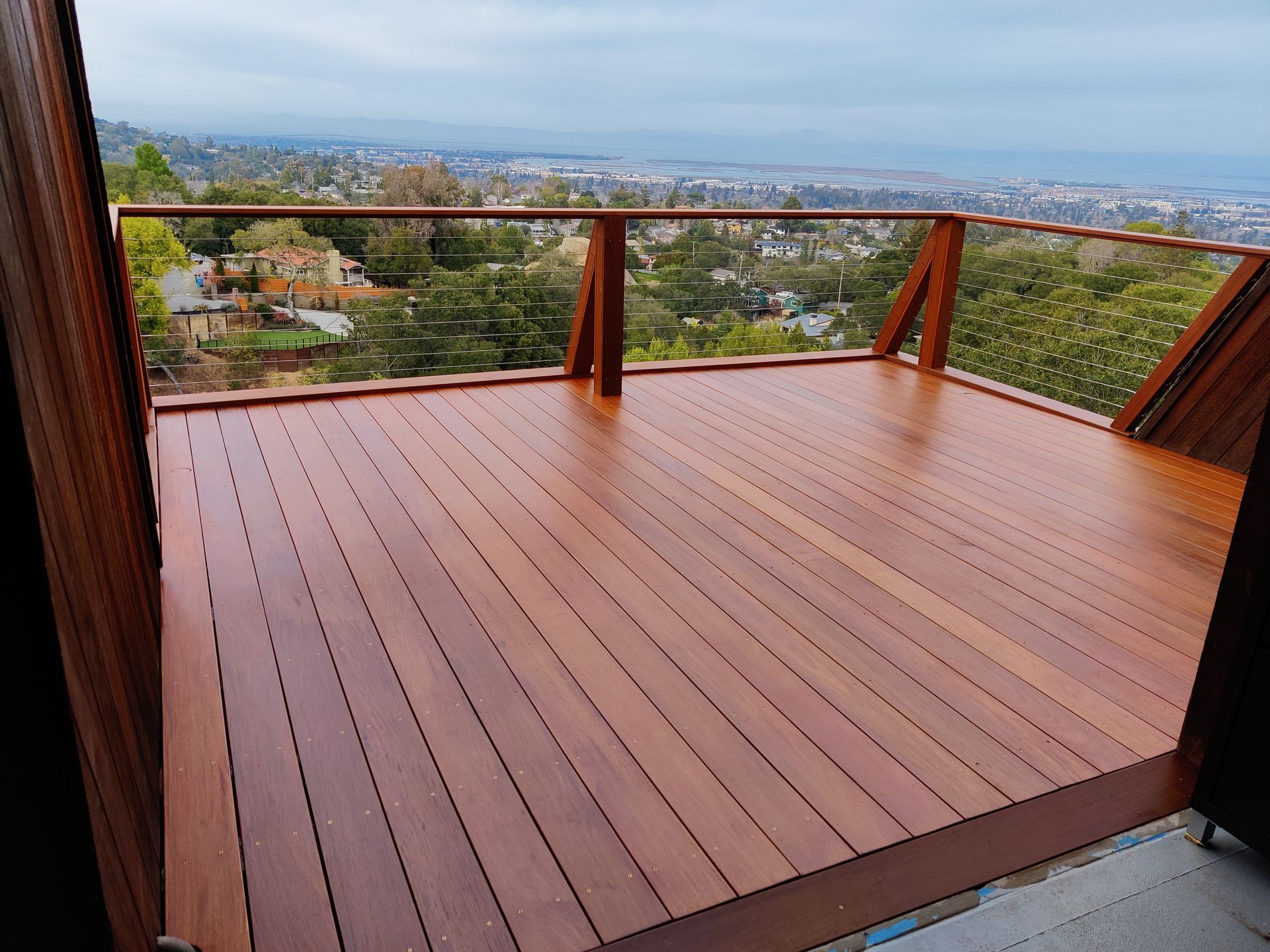 Wooden deck with steps and railing next to a white house with black window frames, surrounded by trees.