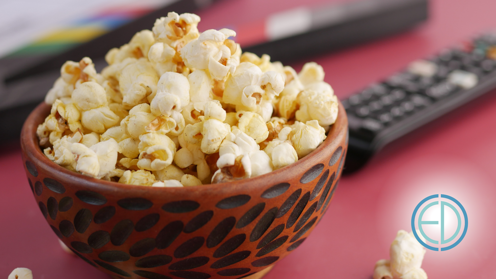 Bowl of popcorn with TV remotes in the background on a pink surface.