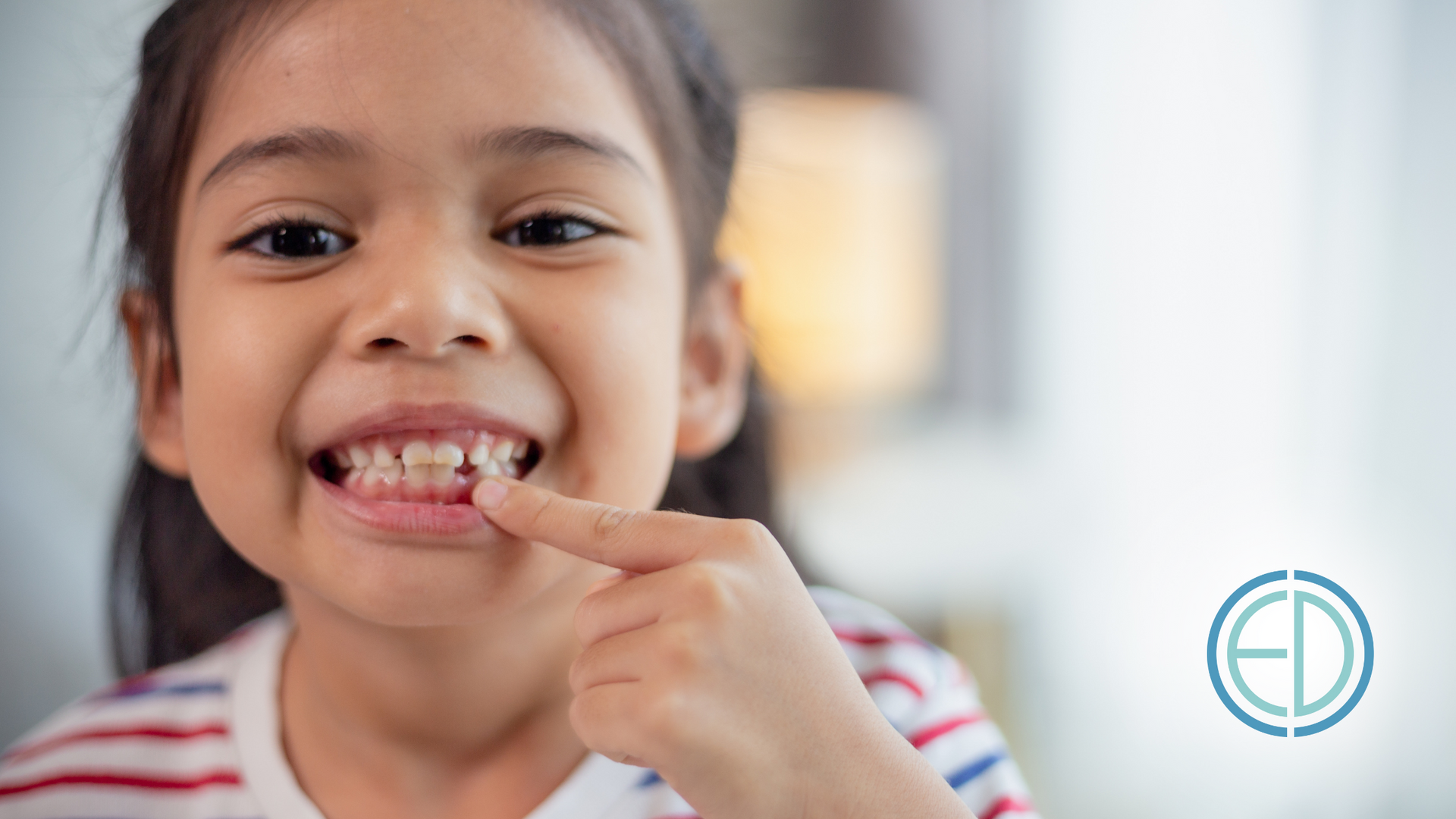 A little girl is pointing at her teeth and smiling.