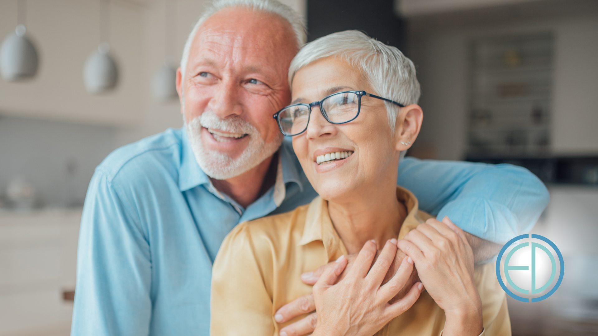 Older couple smiling, embracing indoors. Man has arm around woman, they are looking to the side.
