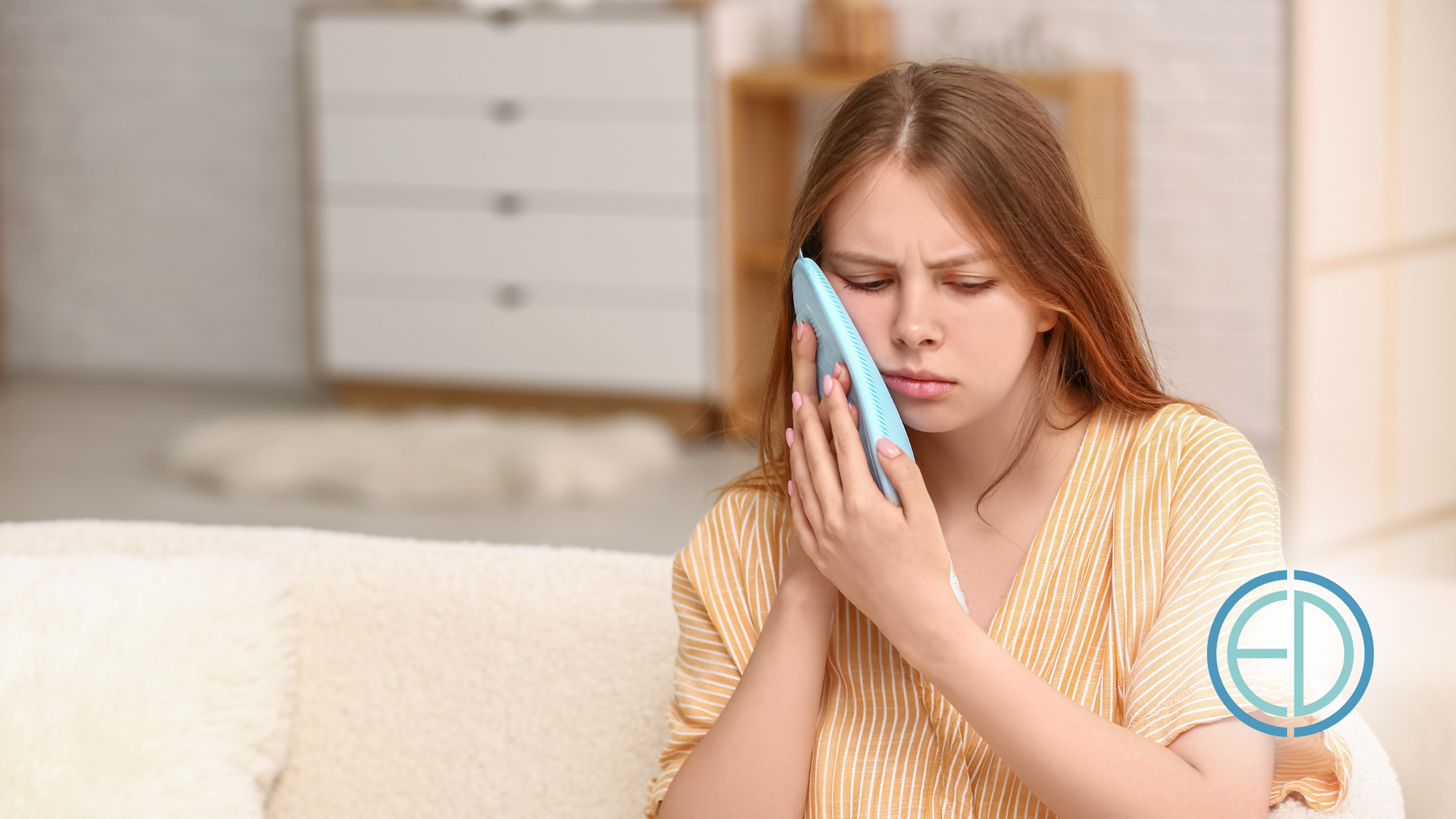 A woman is sitting on a couch holding an ice pack to her face.