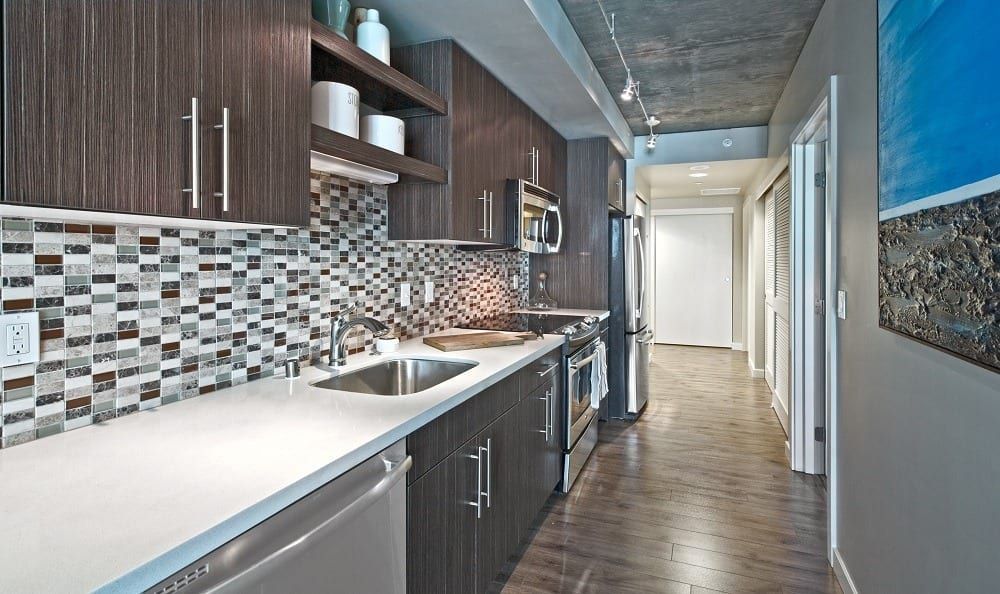 Galley-style apartment kitchen with dark wood cabinets, stainless appliances, and mosaic backsplash.