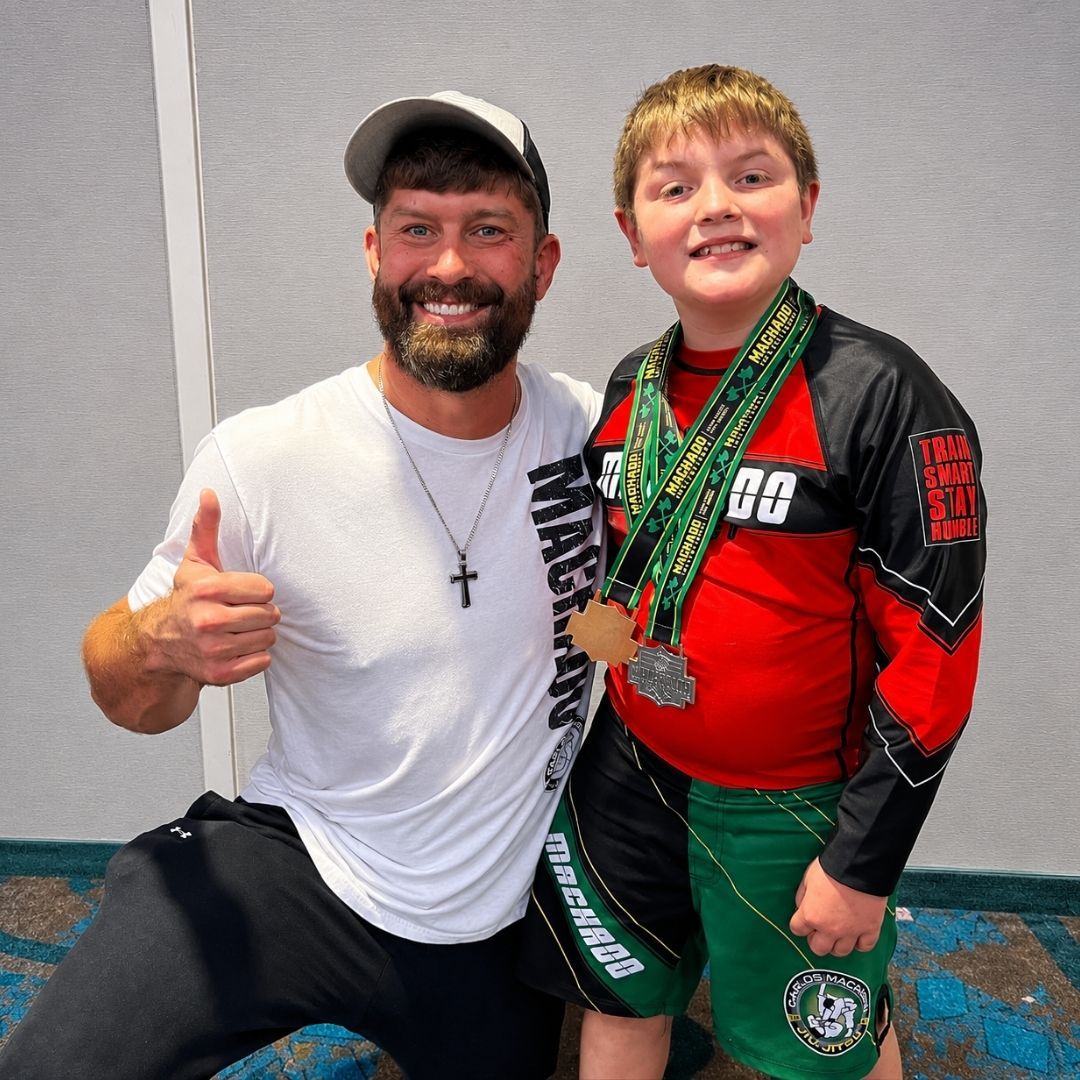 Two boys on a mat practicing Jiu-Jitsu. One boy is on top, giving a thumbs-up.