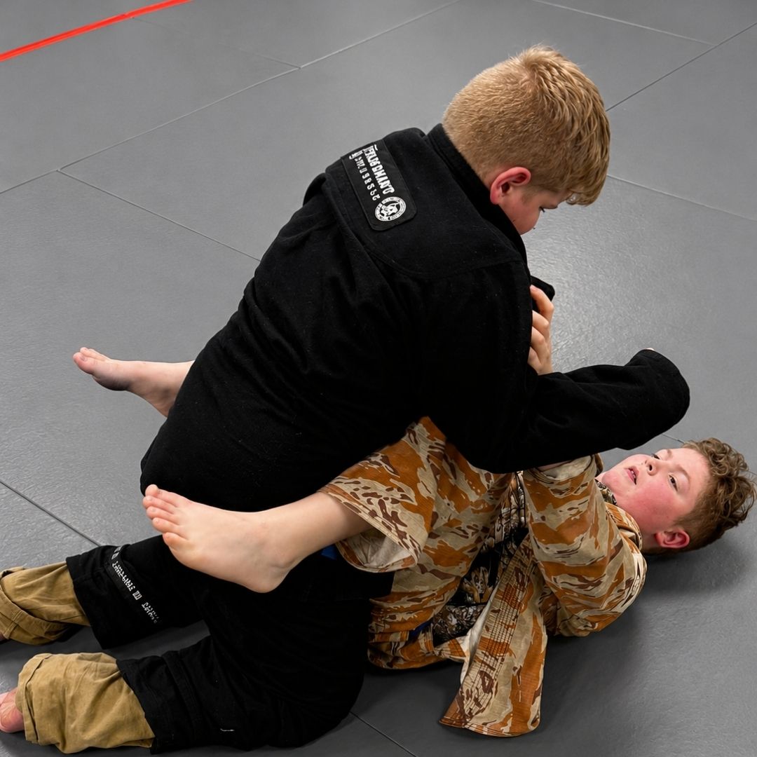 People practicing Brazilian Jiu-Jitsu. One person on the ground, another on top; two others grappling nearby. Black gis, indoor setting.