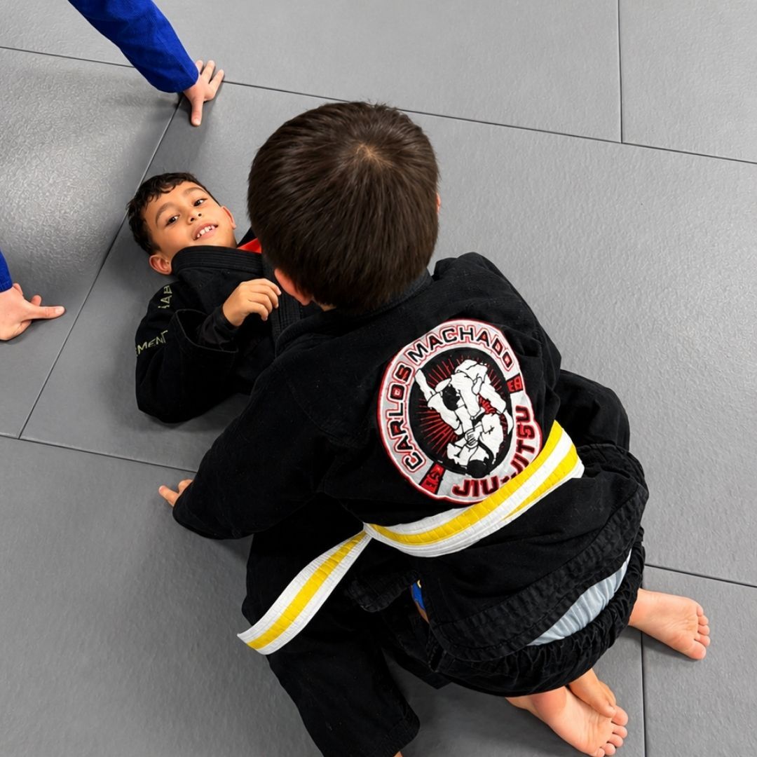 Two children in black and white martial arts uniforms practice a chokehold on a mat. One child smiles.