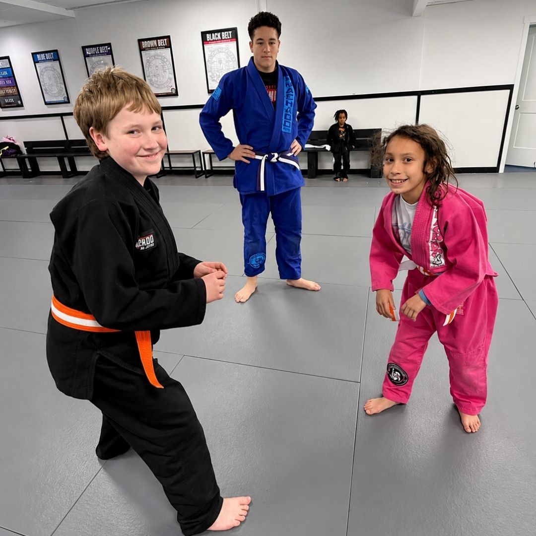 Two children practicing Brazilian Jiu-Jitsu on a mat. One child in white gi has the other in a leg lock. A girl watches.