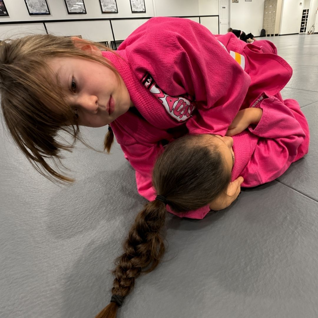 Two people grappling on a mat, one in a black gi, the other in a rash guard, legs entangled.