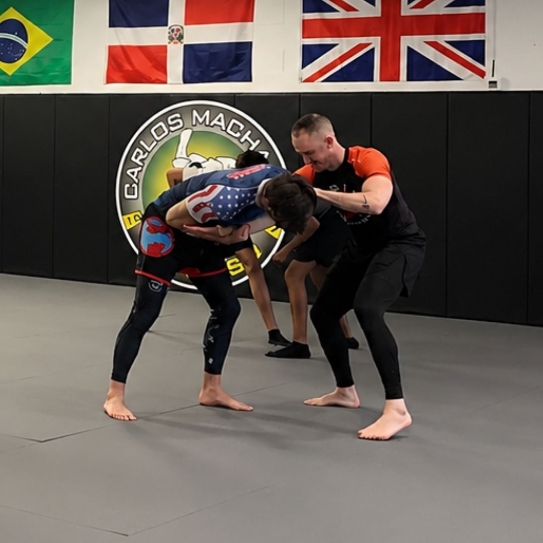 Two men in grappling position on a mat. One embraces the other from behind, both smiling. Gym setting.