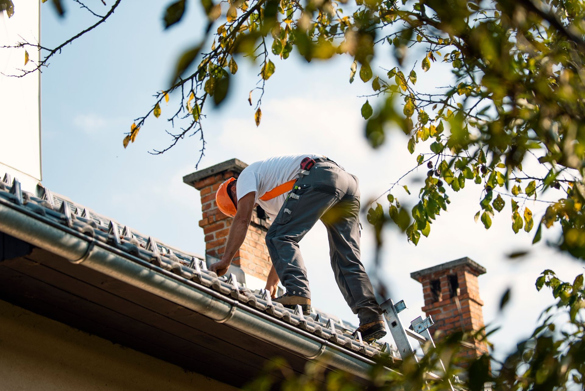Roofer on a ladder, working on a roof near a chimney, wearing a hardhat and safety harness.