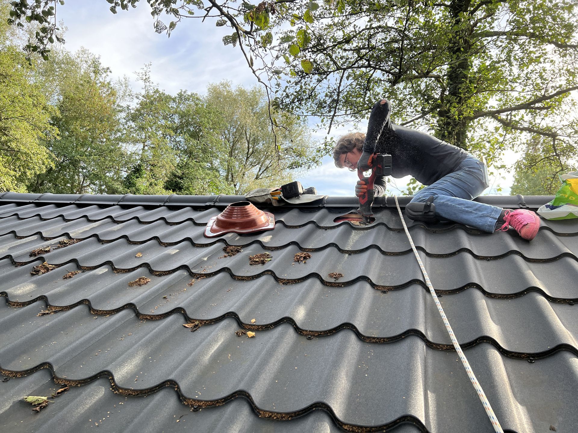 Person on a gray metal roof using a drill. They are wearing jeans, a dark jacket, and pink shoes. Person on a gray metal roof using a drill. They are wearing jeans, a dark jacket, and pink shoes.