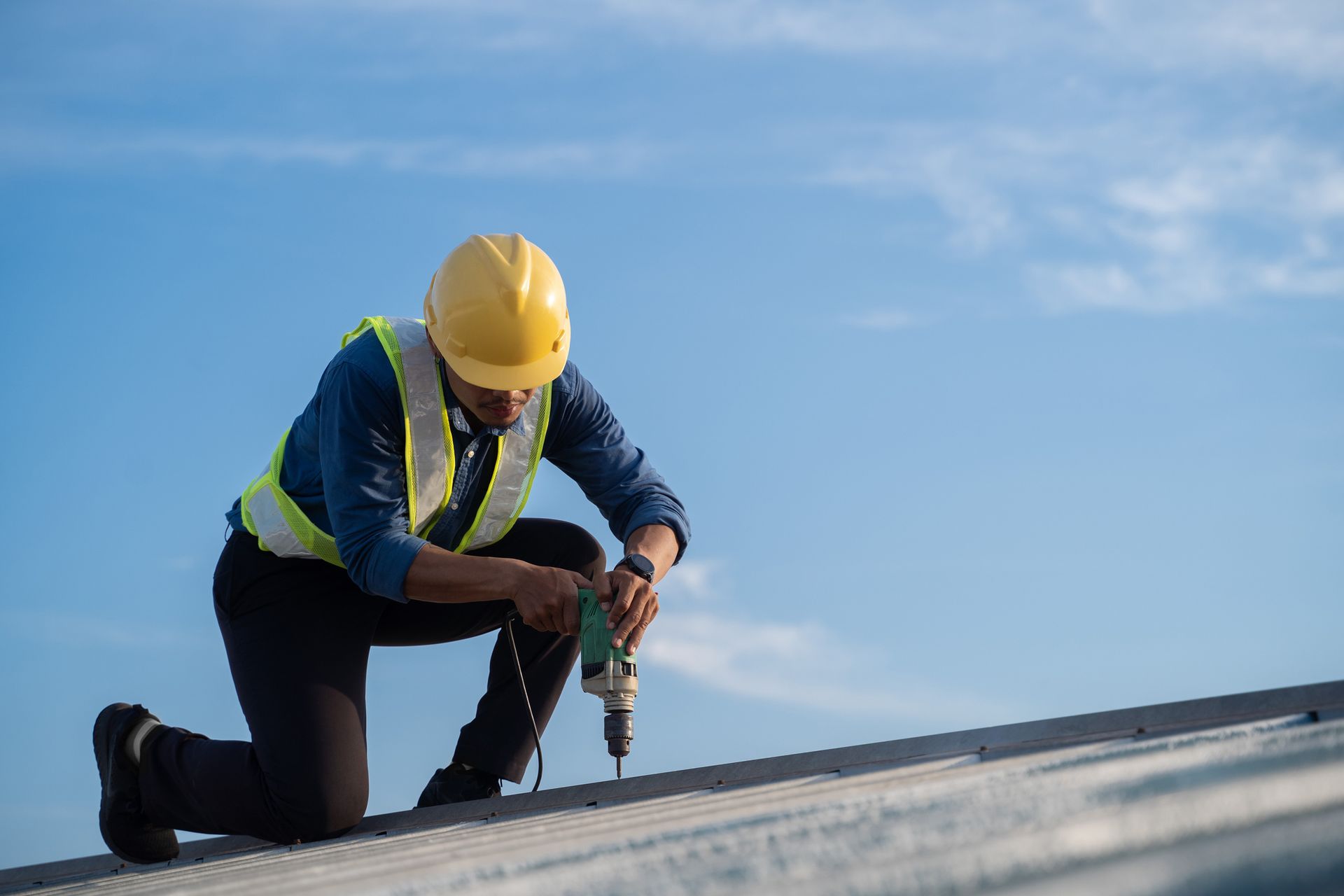 Construction worker kneeling, using a drill on a rooftop under a blue sky. Wearing yellow helmet and safety vest.