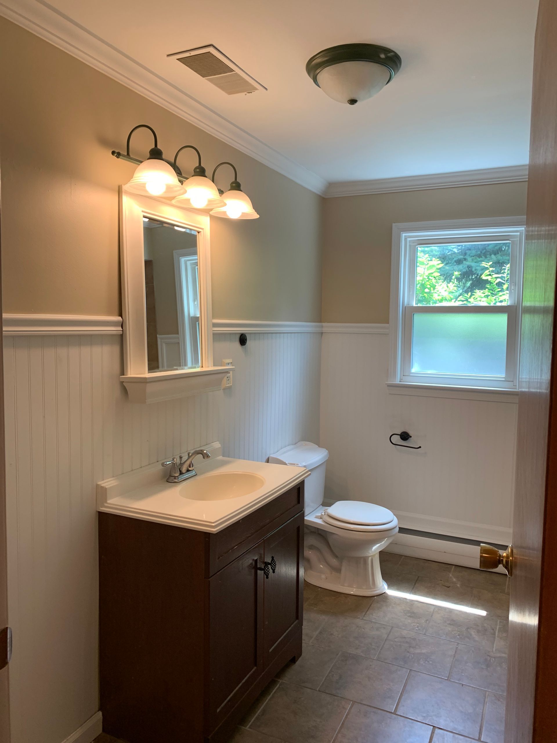A bathroom with a sink , toilet , mirror and window.