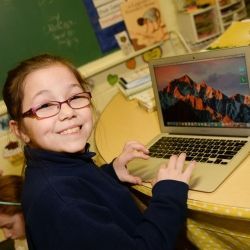 A young girl wearing glasses is using a laptop computer in a classroom.