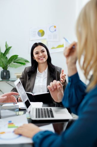 Reunión de negocios: una mujer con blazer sonríe, haciendo un gesto hacia la computadora portátil y sus colegas, con gráficos en el fondo.