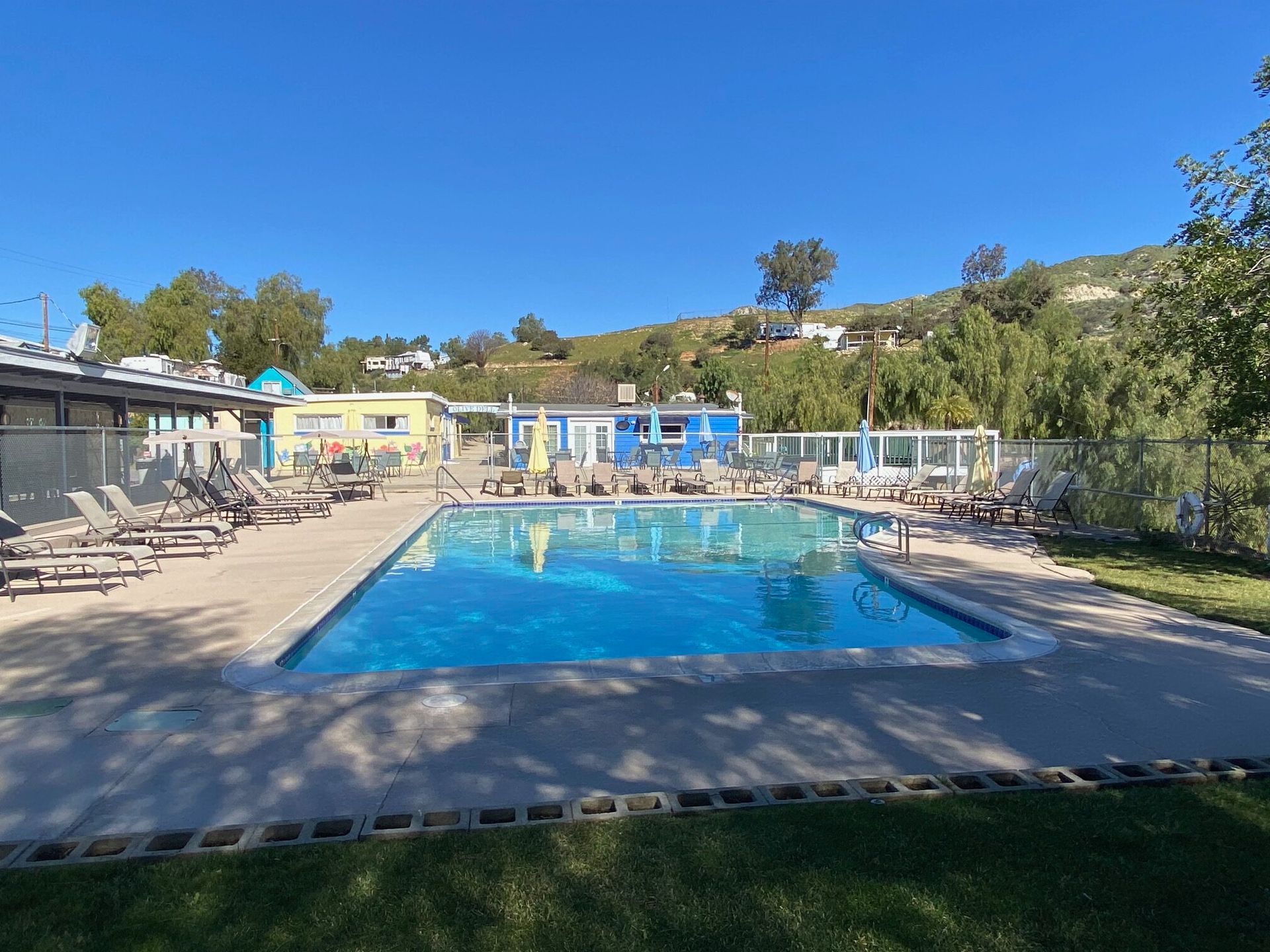 A sunny outdoor swimming pool surrounded by lounge chairs, with small buildings and hills in the background.