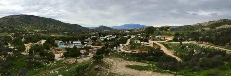 A panoramic view of a small residential neighborhood nestled in a valley among rolling, green, tree-covered hills.