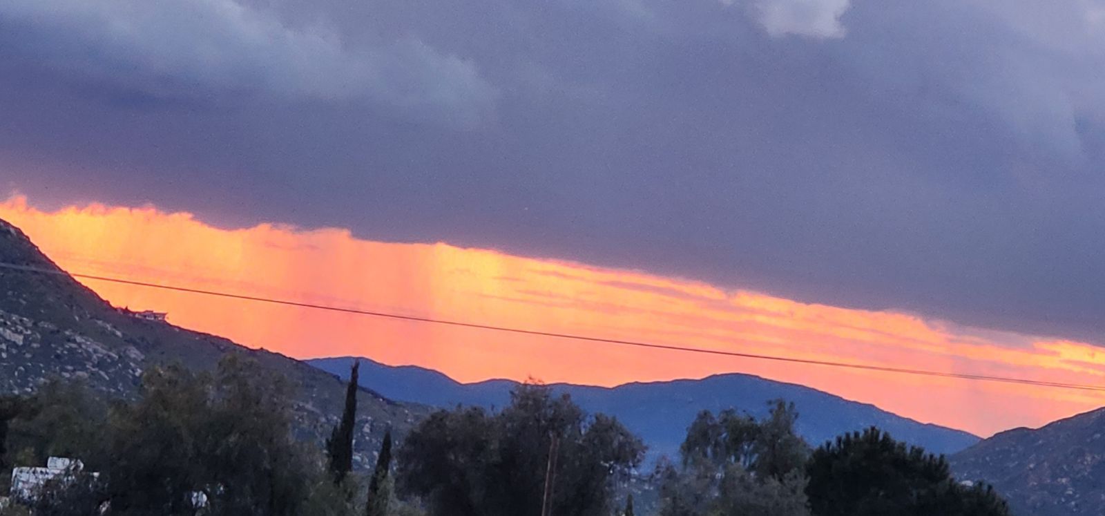 A vibrant sunset glows behind dark, stormy clouds above silhouetted mountains and trees, with a power line crossing.