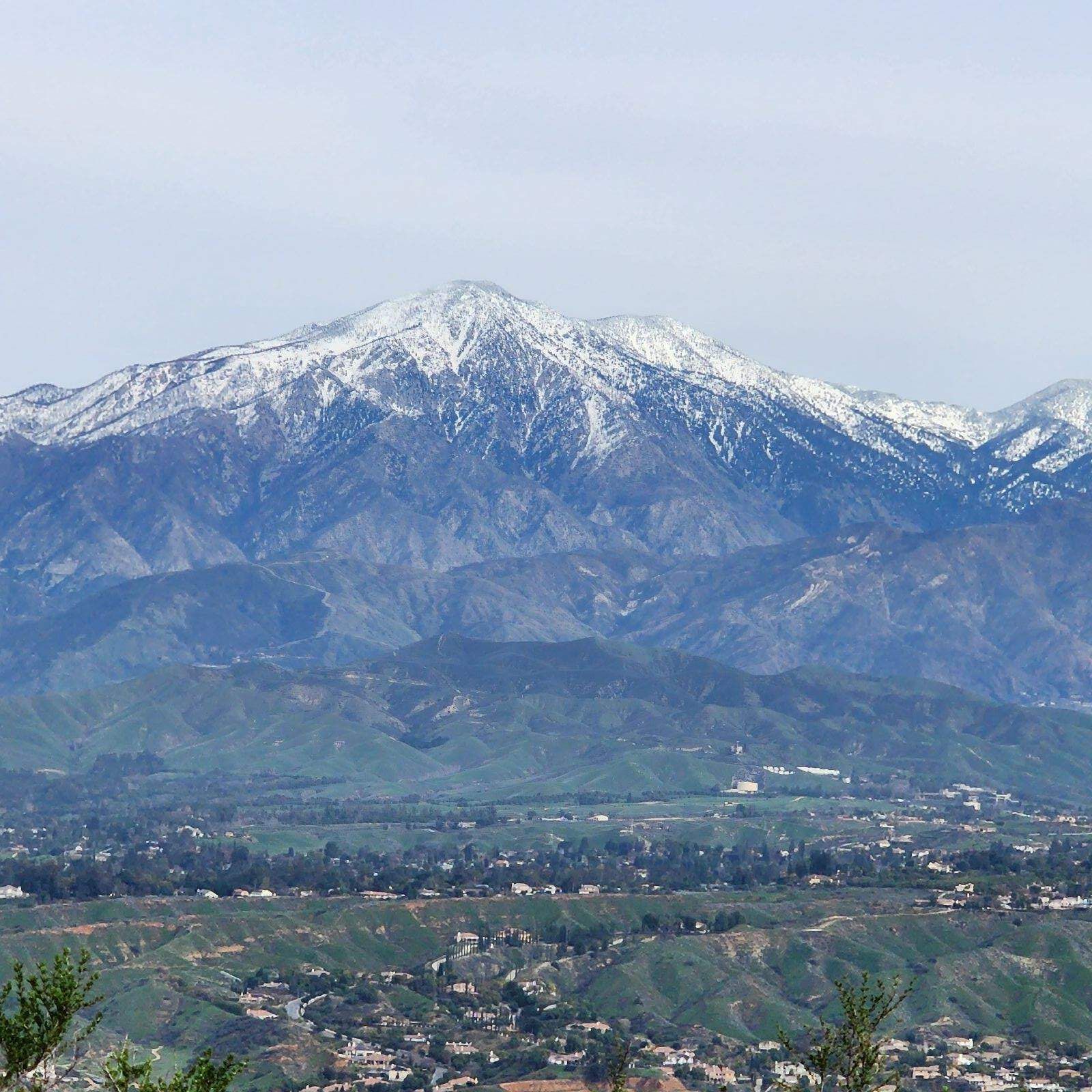 A snow-capped mountain peak towers over rolling green hills and a residential valley landscape under a pale sky.