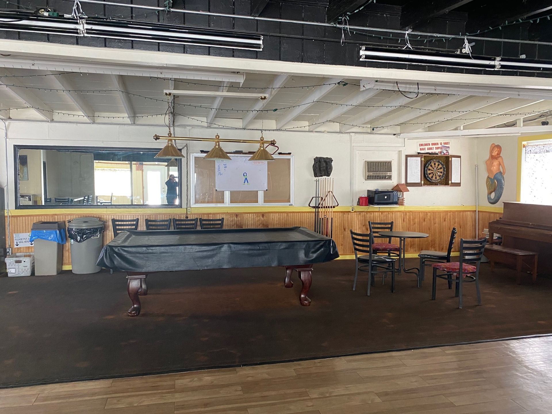 A pool table covered with a black cloth stands in a room with wood-paneled walls, chairs, and a dartboard.