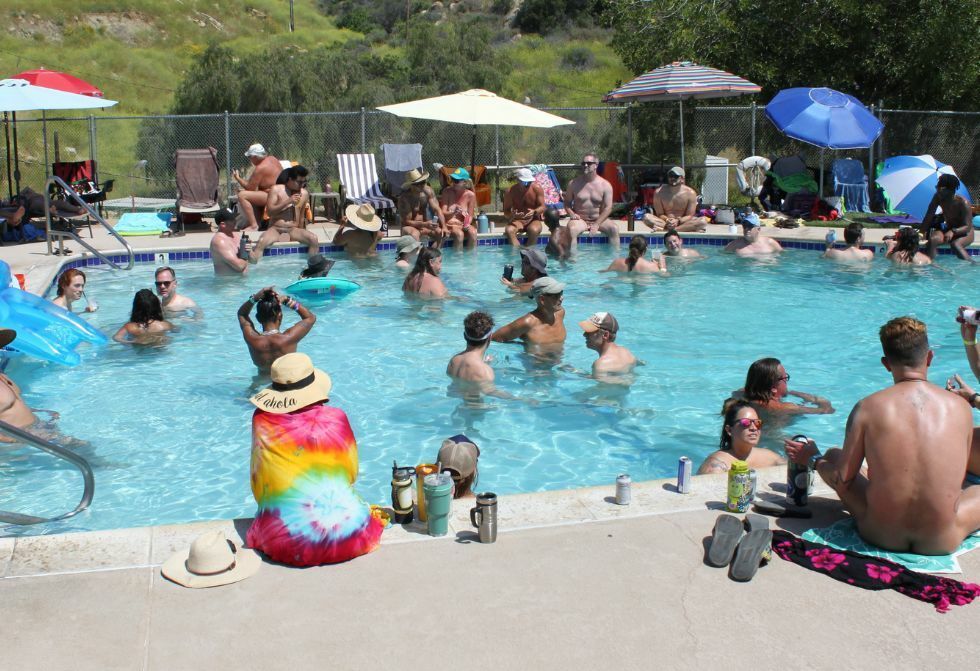 Many people swim and relax in a sunny outdoor swimming pool surrounded by lounge chairs, umbrellas, and grassy hills.