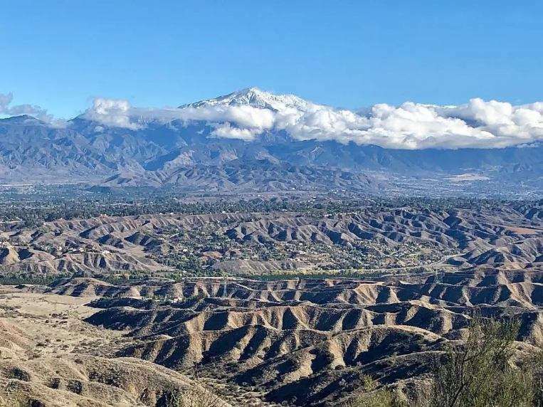 A snow-capped mountain range rises behind rugged, sunlit desert badlands under a clear blue sky.