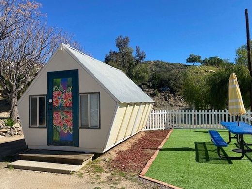 A small A-frame cabin with a floral-painted door, next to a picnic table and an umbrella on a patch of artificial grass.