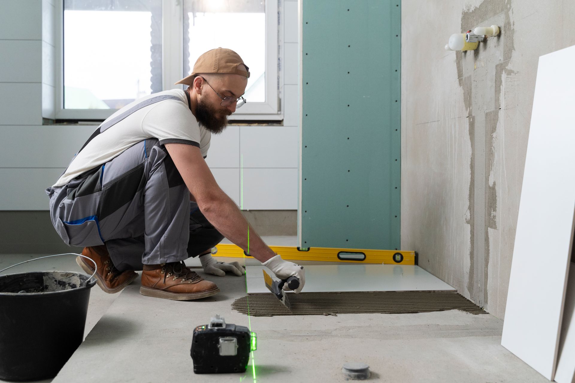 Man tiling a floor in a bathroom, using a trowel and level. Green laser level on floor.