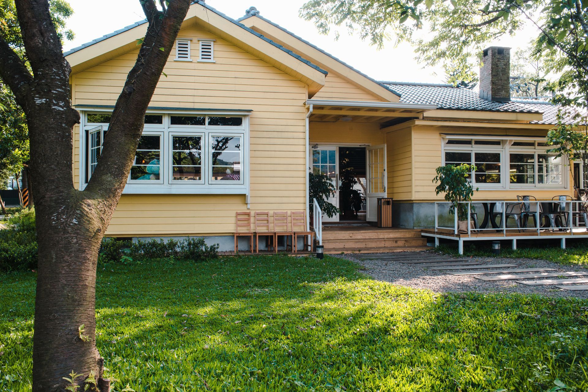 Yellow cottage with white-framed windows, a porch, and green lawn; a tree in the foreground.